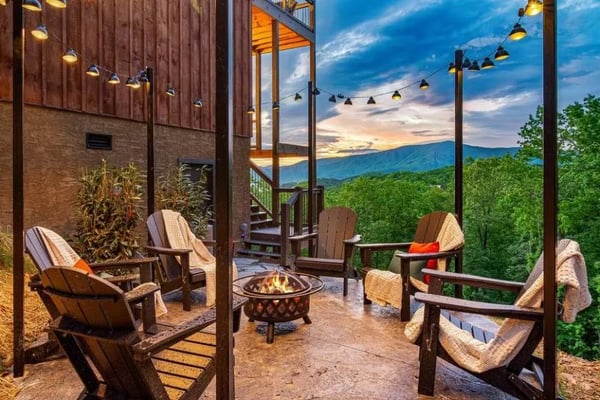 Covered patio with Adirondack chairs, fire pit, and mountain sunset view at dusk with string lights
