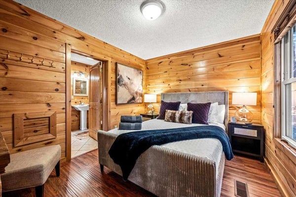 Wooden cabin bedroom with queen bed, dark bedding, nightstands with lamps, and ensuite bathroom visible through open door