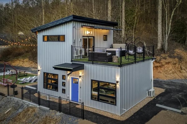 Modern two-story metal cabin with blue door, upper deck, and black railing surrounded by bare trees and recreational grounds.
