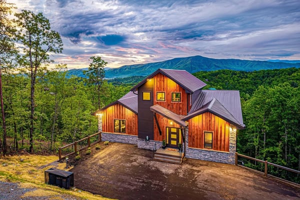 Modern cabin with red wood siding and metal roof overlooking forested mountains at dusk
