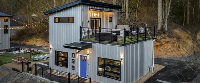 Modern two-story metal cabin with blue door, upper deck, and surrounding recreational grounds in wooded setting.
