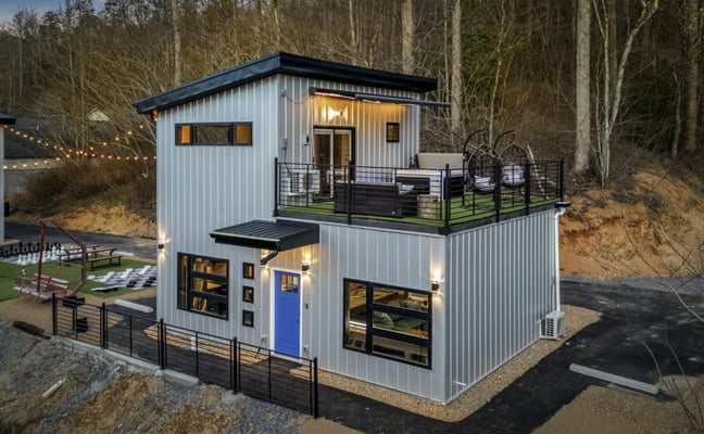 Modern two-story metal cabin with blue door, upper deck, and black metal railing surrounded by bare trees and recreational grounds.